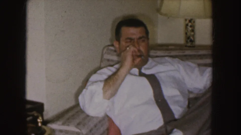 Nebraska 1955 frame of a man in shirt sleeves and suspenders reclining indoors while holding a cigar near his mouth.
