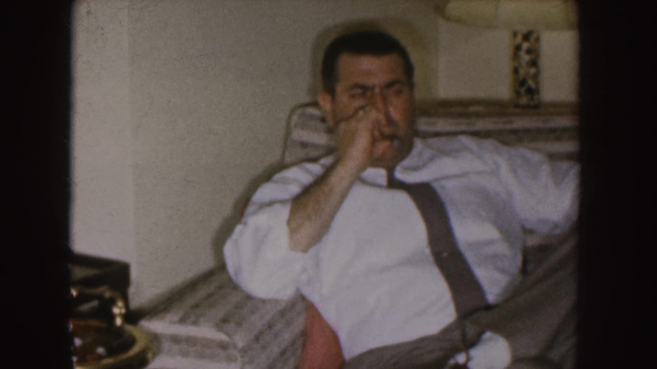 In 1955 Nebraska home-movie footage, a man in a white shirt and suspenders sits in a patterned armchair while smoking.