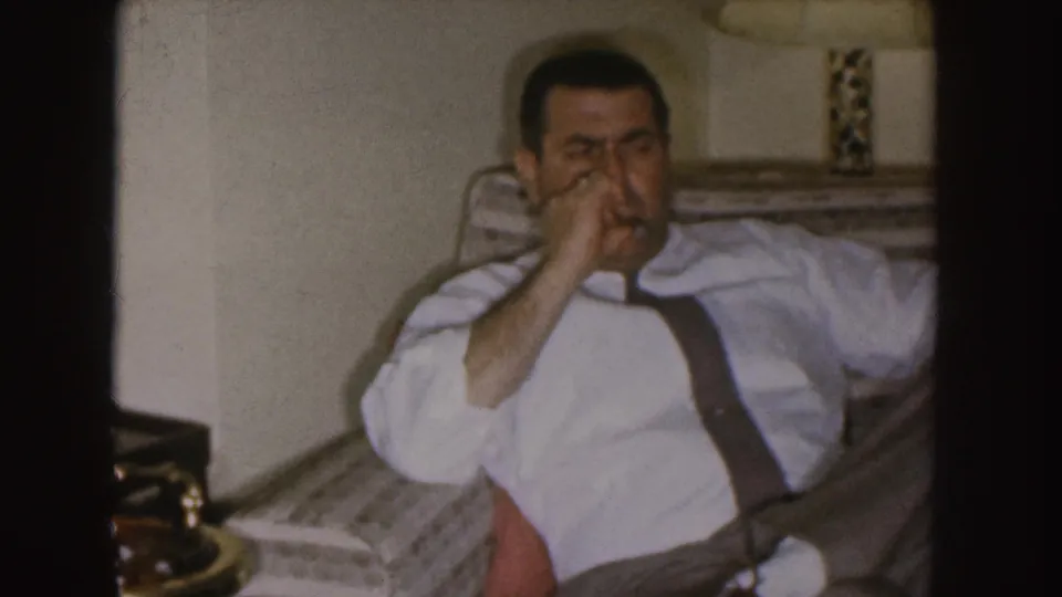 In 1955 Nebraska home-movie footage, a man in a white shirt and suspenders sits in a patterned armchair while smoking.
