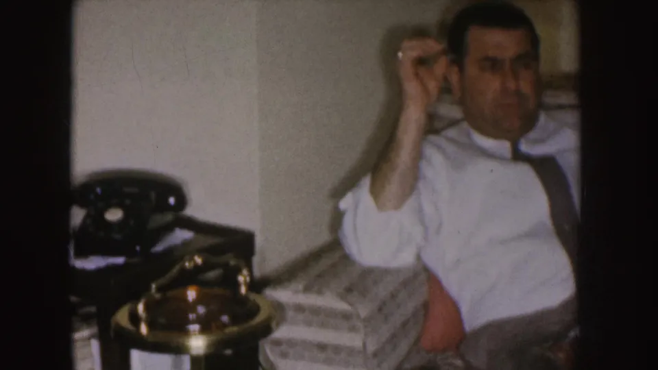 The seated man remains in the armchair as a black rotary telephone and brass-toned lamp appear on the nearby table.