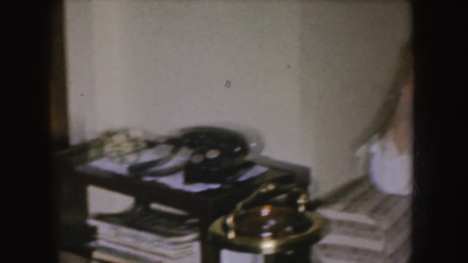 The handheld view shifts toward the side table, showing a black rotary telephone, papers or magazines, and a brass-toned lamp.