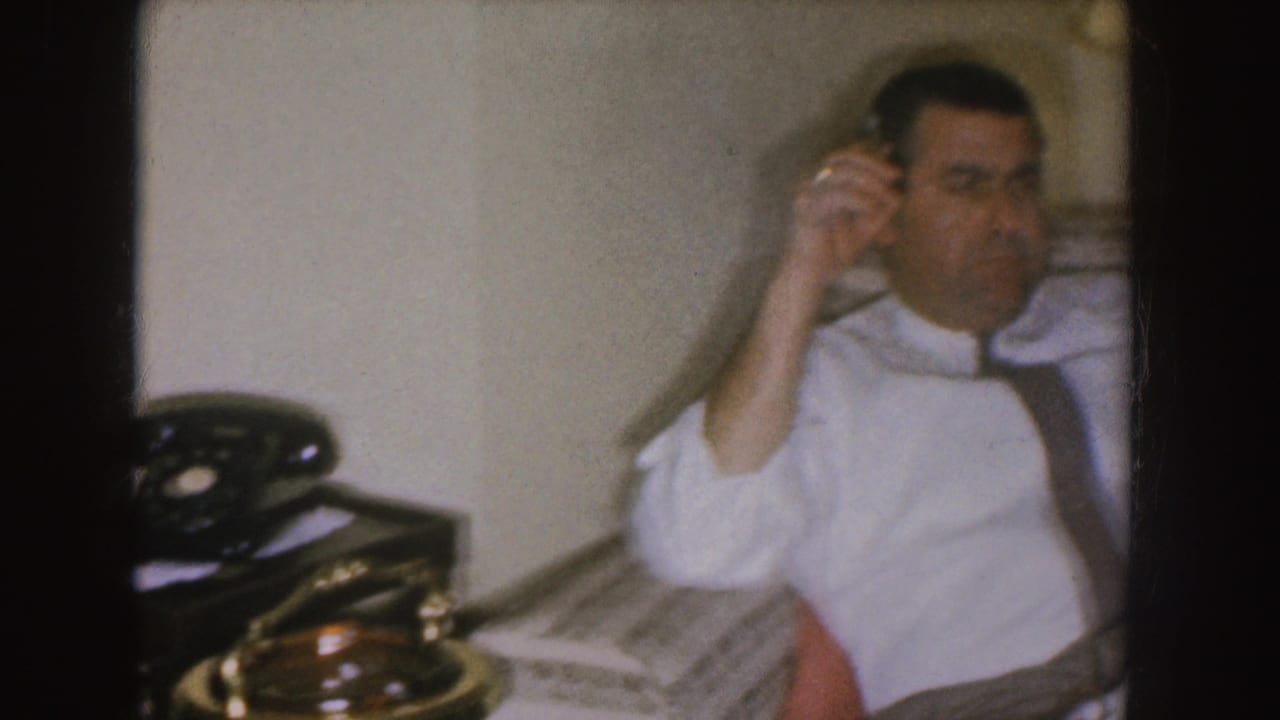 Man seated on a patterned couch holding a cigarette near a rotary telephone.