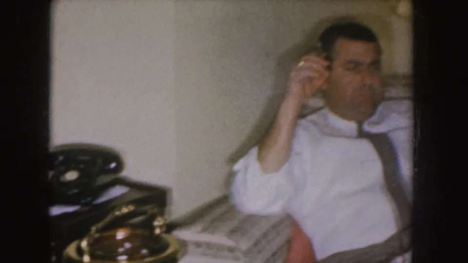 In a Nebraska 1955 home interior, a man in suspenders sits on a patterned couch with a cigarette beside a rotary telephone.