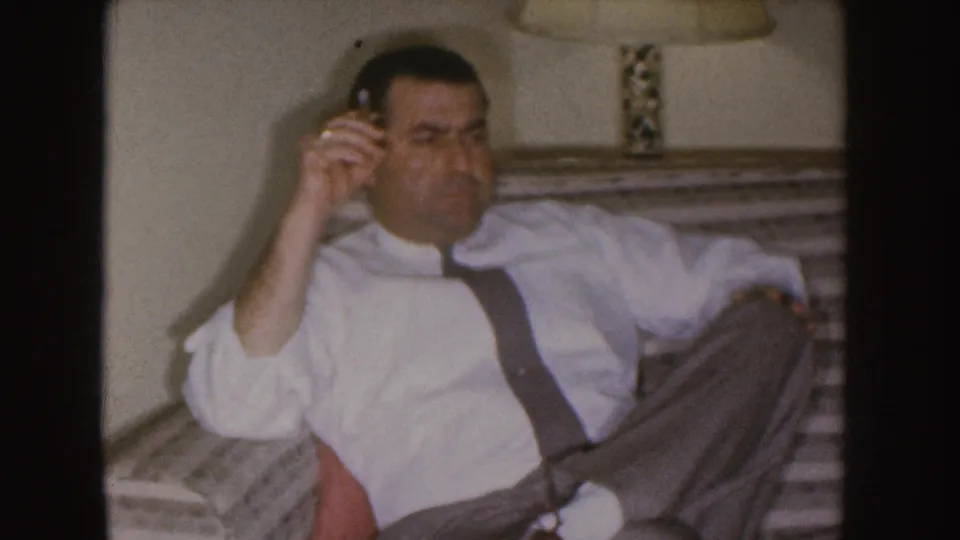 A 1955 Nebraska home-movie frame shows the man seated on the couch, cigarette in hand, facing toward the camera.