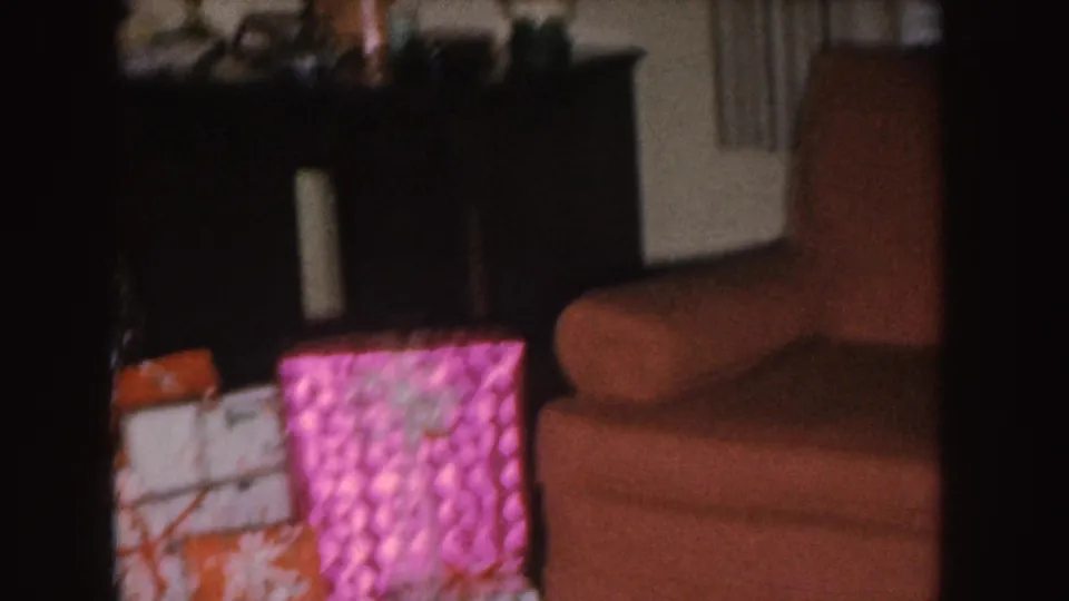 1955 Nebraska home-movie frame with wrapped Christmas gifts beside a brown couch and dark furniture in a private interior.