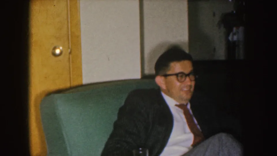 1955 Nebraska home-movie frame showing a smiling man in a suit and glasses seated on a green couch in a private living room.