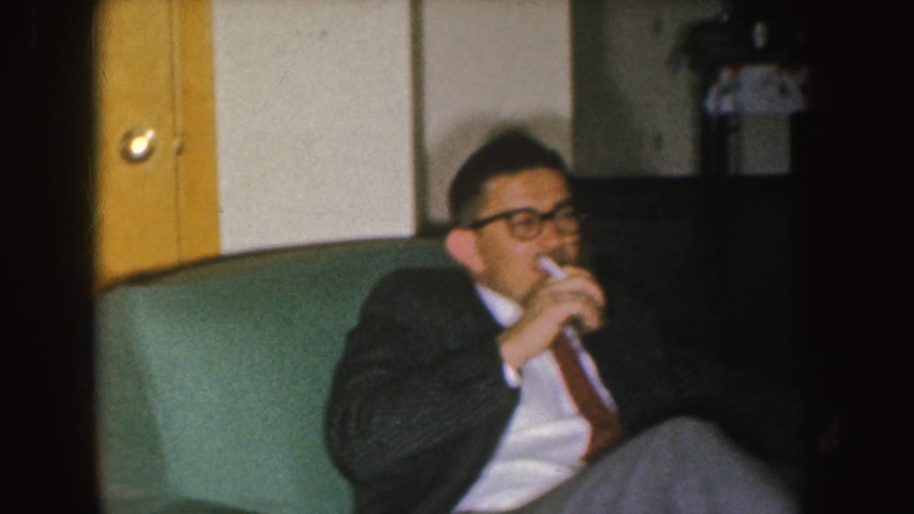 In 1955 Nebraska home-movie footage, a man in glasses sits on a green couch with a drink near a wooden door.
