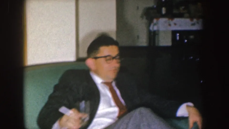The seated man lowers his drink while framed against a green couch and household furnishings in a Nebraska home.