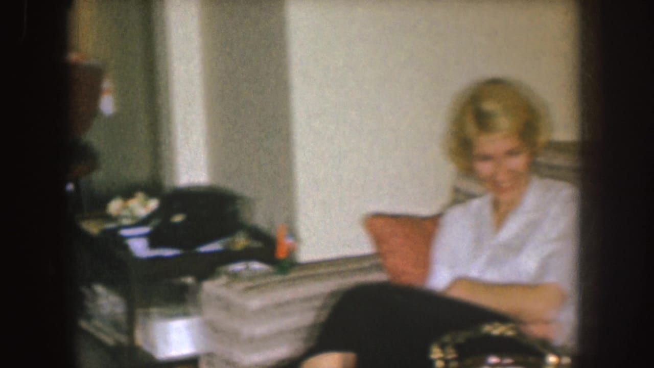 In a 1955 Nebraska home-movie frame, a blonde woman sits smiling on a striped couch beside a side table and cushion.