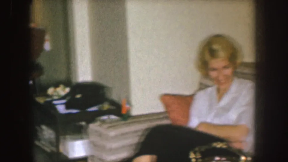 In a 1955 Nebraska home-movie frame, a blonde woman sits smiling on a striped couch beside a side table and cushion.
