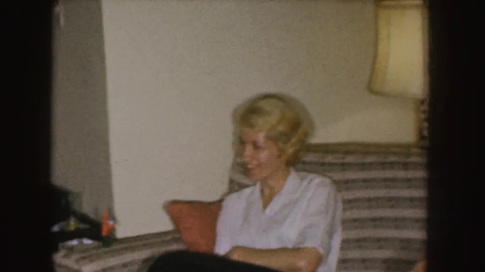 The woman smiles from a striped couch in a Nebraska living room, with a shaded lamp and orange cushion visible nearby.