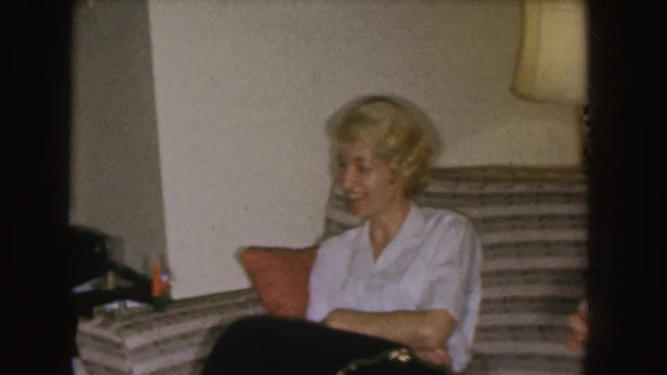 A blonde woman in a white blouse sits on a striped couch, smiling toward someone outside the left edge of the frame.