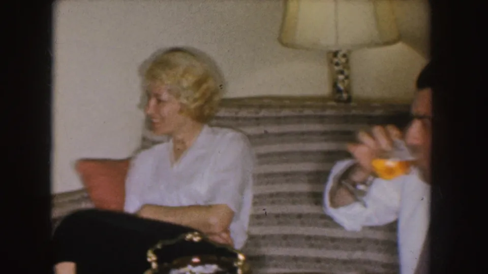 The woman laughs on a striped couch while a partially visible man at the right holds a drink in the living room.