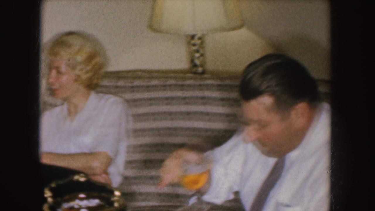 In a 1955 Nebraska home-movie frame, a blonde woman sits on a striped couch as a man beside her sips from a glass.