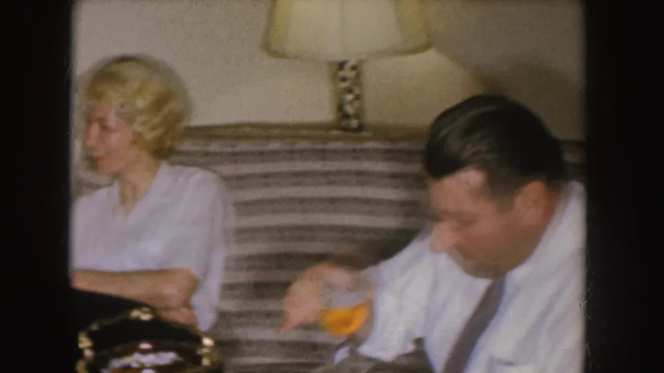In a 1955 Nebraska home-movie frame, a blonde woman sits on a striped couch as a man beside her sips from a glass.