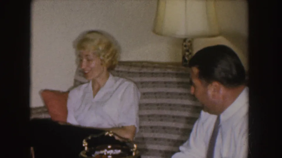 The woman smiles from a striped couch in a Nebraska living room while the man sits nearby, partly turned toward her.