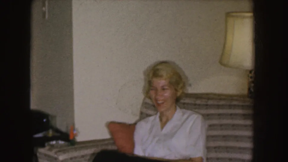 A blonde woman in a white blouse smiles on a striped couch in the 1955 Nebraska living-room scene.
