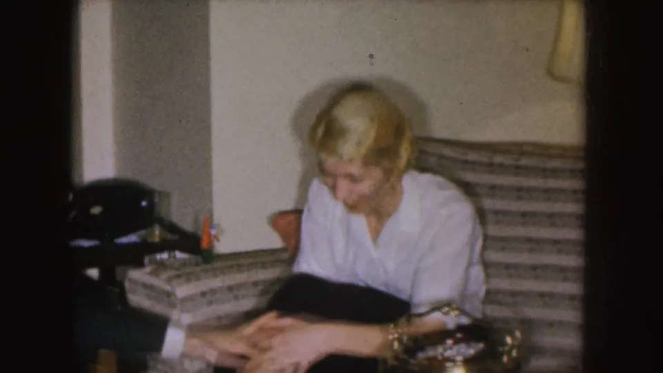 In a Nebraska 1955 home-movie frame, a blonde woman in a white blouse and dark skirt sits on a striped couch near a side table.