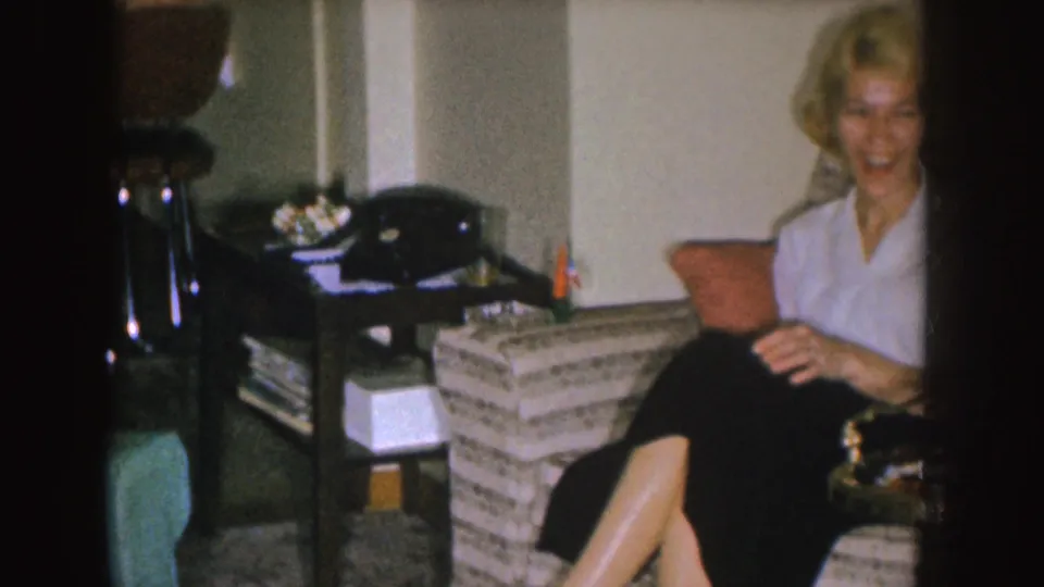 A smiling woman in a white blouse and dark skirt sits in a mid-century Nebraska living room near a striped couch and black rotary phone.