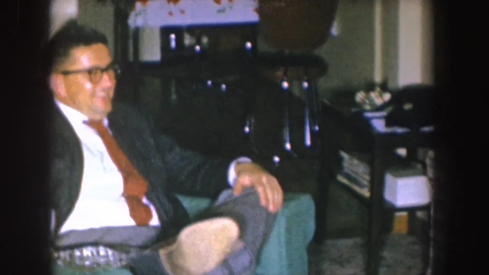 A man wearing glasses, a dark suit, white shirt, and reddish tie sits in a green armchair in a Nebraska home interior.