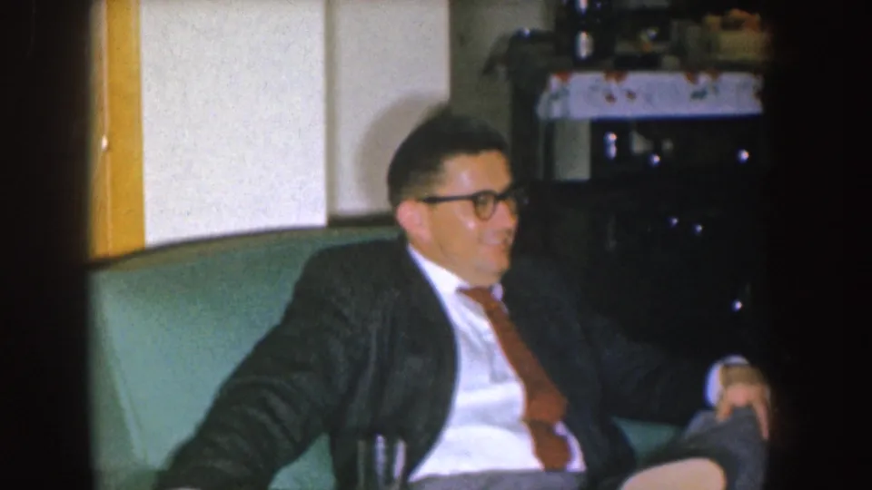 A suited man with glasses reclines in a green armchair, with a side table and living-room furnishings visible behind him.