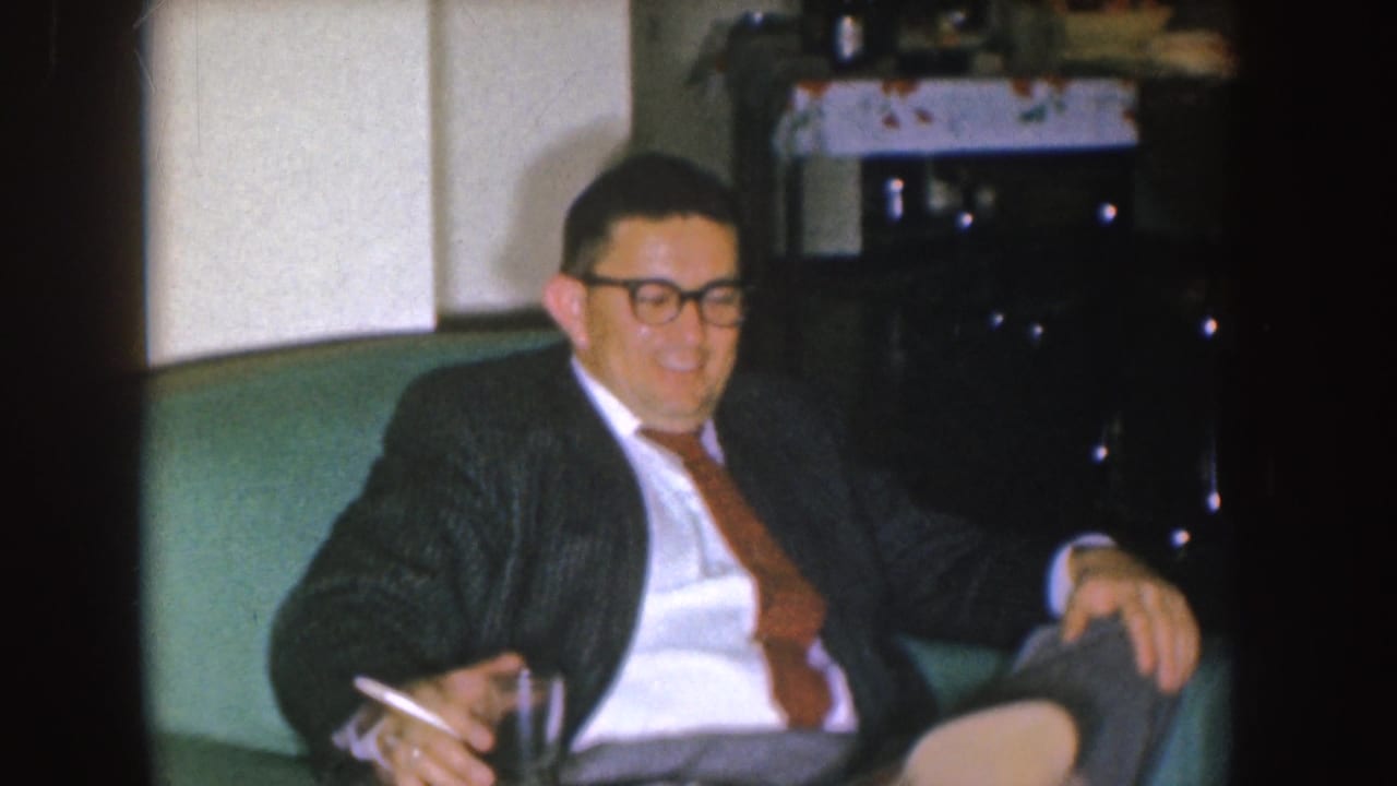 In a 1955 Nebraska home-movie frame, a suited man with glasses sits on a green couch holding a cigarette.