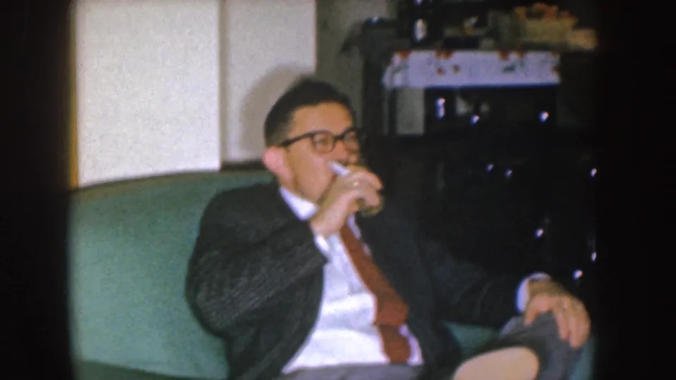 In a Nebraska 1955 home interior, a suited man with glasses drinks from a glass while seated on a green couch.