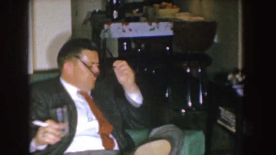 A suited man in a 1955 Nebraska living room holds a glass and cigarette while seated on a green couch.