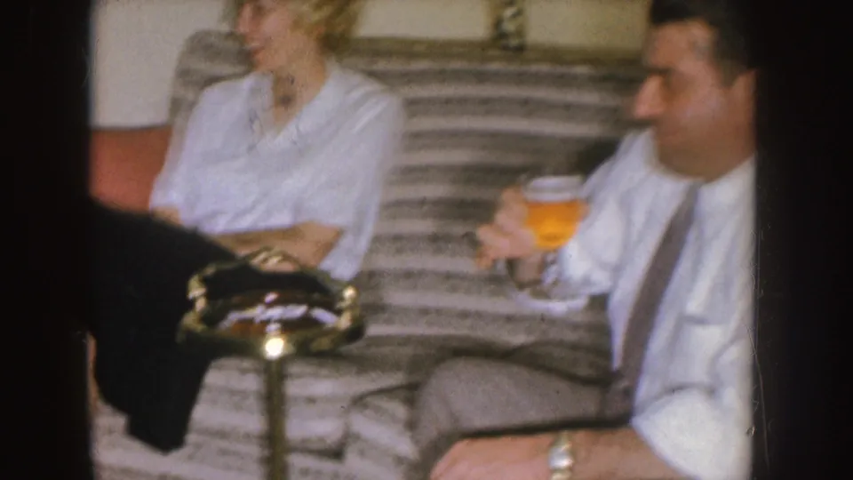 In the Nebraska home-movie frame, a woman and man sit on a striped sofa while the man holds a glass of beer.