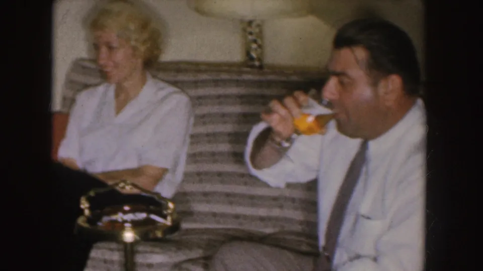 The man drinks from a beer glass beside the seated woman in a 1955 Nebraska living-room home movie.
