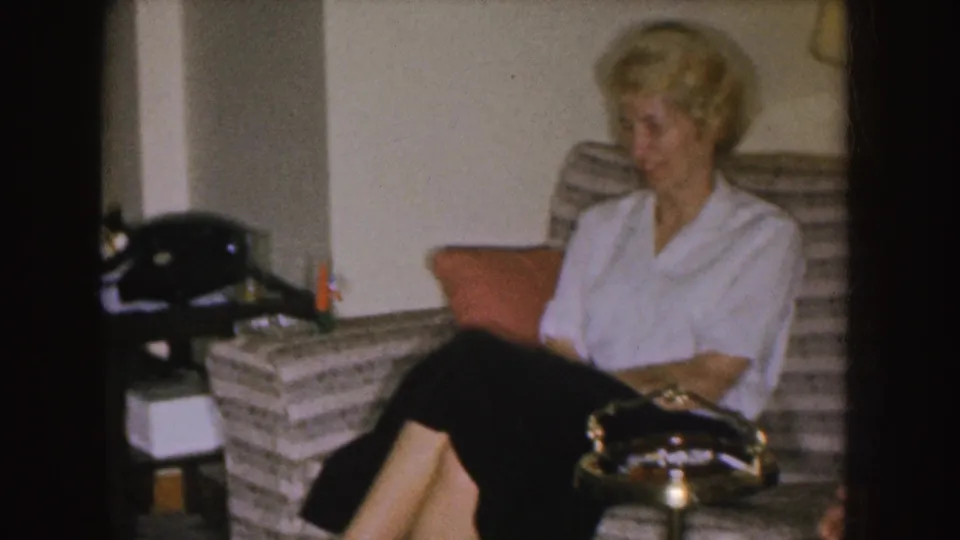 A woman sits on the striped sofa in the Nebraska home interior, with a rotary telephone visible on the side table.