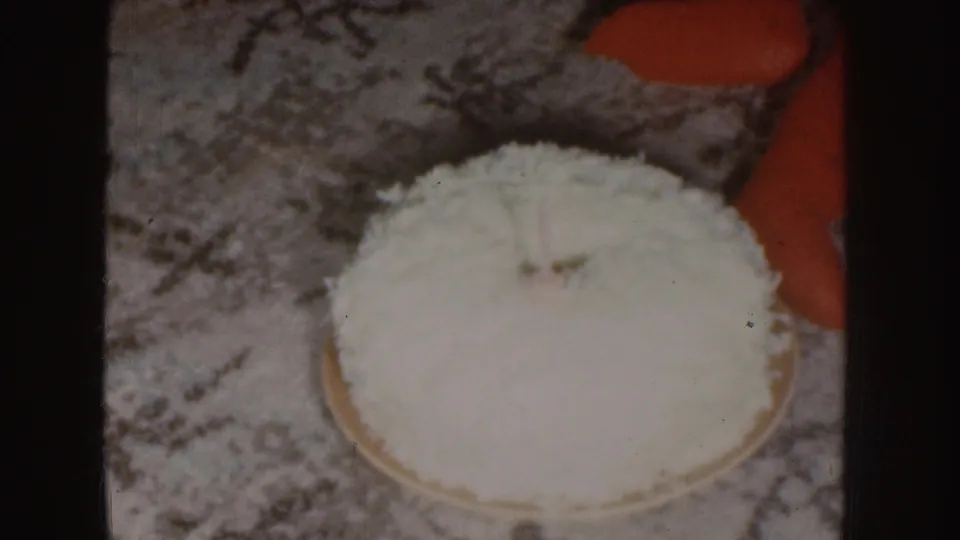 1955 Nebraska 8mm frame showing a round coconut-covered cake on patterned carpet near a child's red sleeve or clothing.