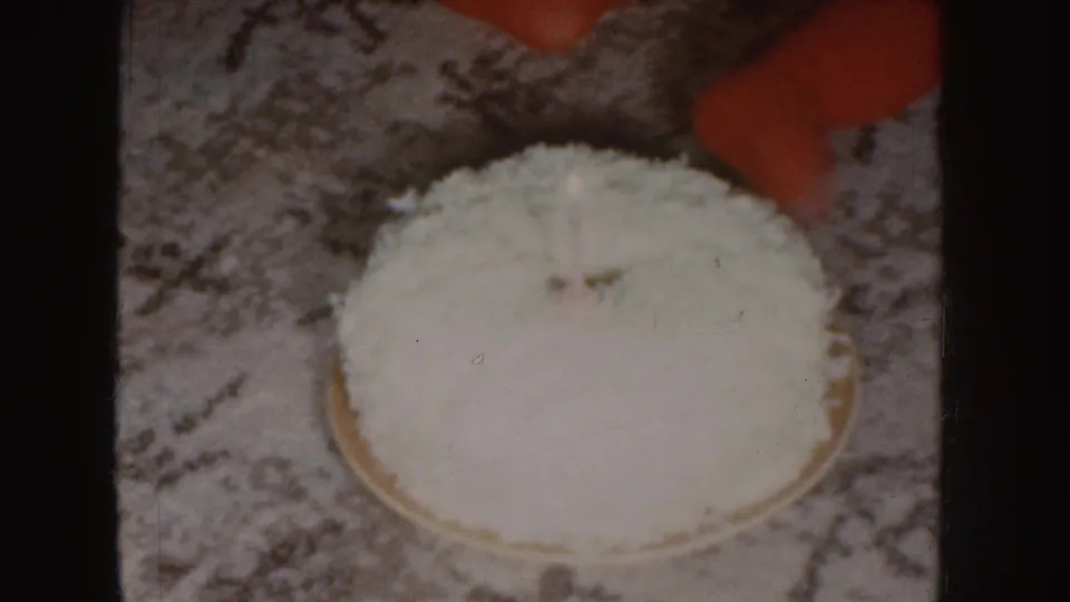 Overhead 1955 Nebraska 8mm view of a white coconut cake on patterned carpet with a child's red clothing partly in frame.