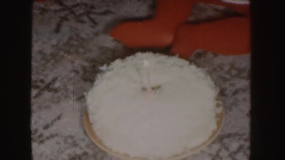 1955 Nebraska home-movie frame of a coconut birthday cake on patterned carpet, with a child's red outfit partly visible behind it.