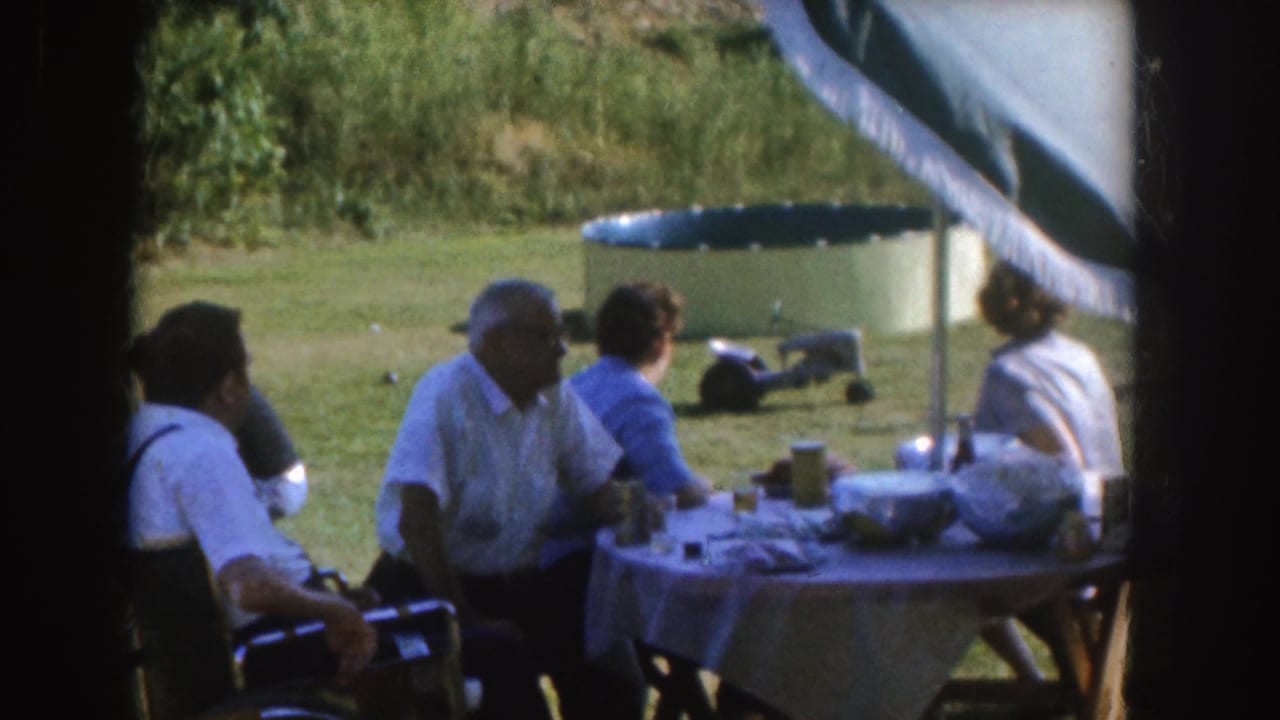 1955 Nebraska home-movie frame showing adults gathered around a covered outdoor table in a grassy yard.