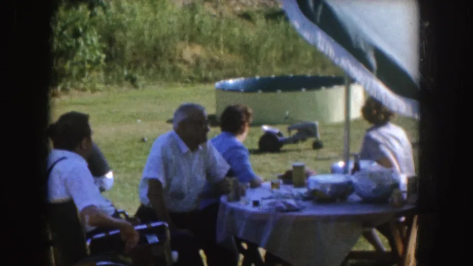 1955 Nebraska home-movie frame showing adults gathered around a covered outdoor table in a grassy yard.