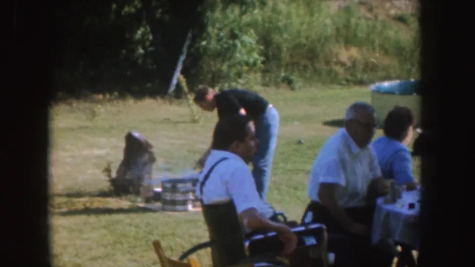 1955 Nebraska home-movie frame showing a backyard meal with seated adults and a grill setup behind them.