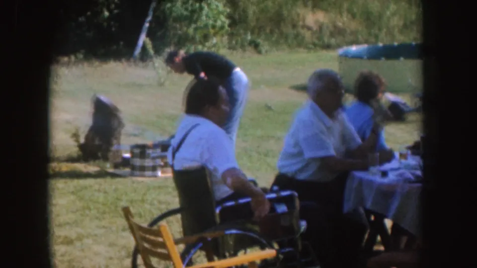 1955 Nebraska frame of adults at a backyard barbecue table, with a wheelchair user and grill visible in the scene.