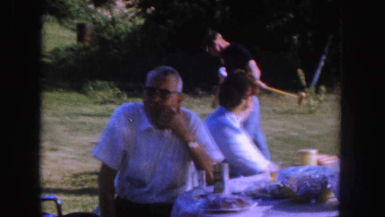 Adult men in white shirts sit at an outdoor table in a grassy yard.