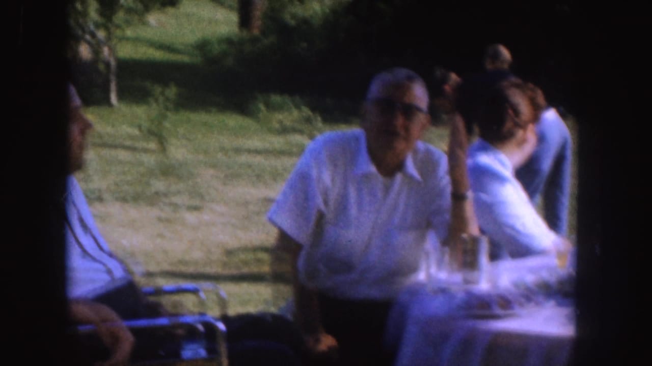 1955 Nebraska frame with an older man wearing glasses seated by a table as another adult man turns in the yard setting.