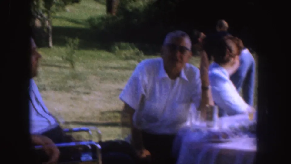 1955 Nebraska frame with an older man wearing glasses seated by a table as another adult man turns in the yard setting.