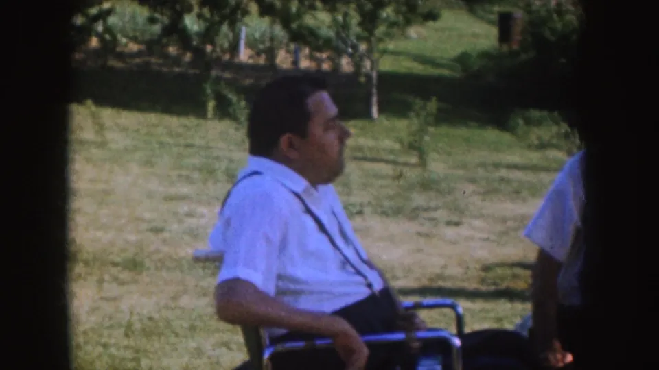 1955 Nebraska home-movie frame showing a man in a wheelchair seated in profile in a grassy yard near other adults.