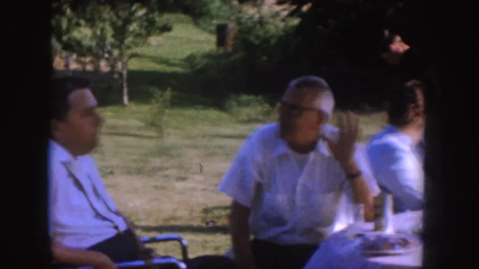 1955 Nebraska frame showing an older man with glasses gesturing during conversation at a cloth-covered table in a yard.