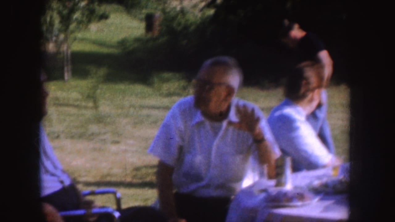 Older man in a white shirt sits at a picnic table while another adult sits beside him outdoors.