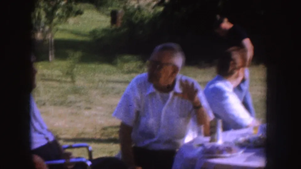 1955 Nebraska home-movie frame showing an older man in glasses at a picnic table, with another seated adult and a person standing in the yard.