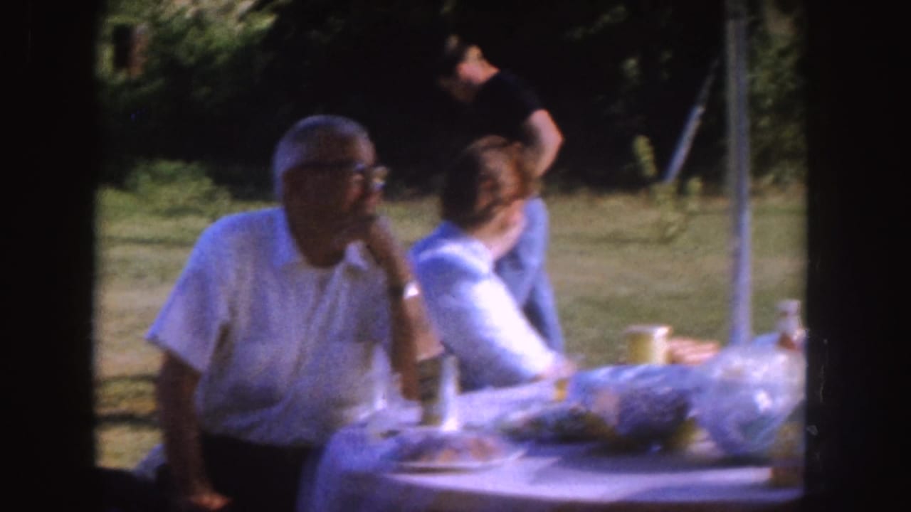 Older man at a picnic table rests his hand near his face as another adult turns away.