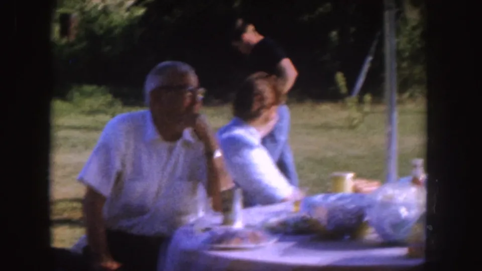 1955 Nebraska backyard picnic frame with an older man in glasses seated at the table and another adult looking to the side.