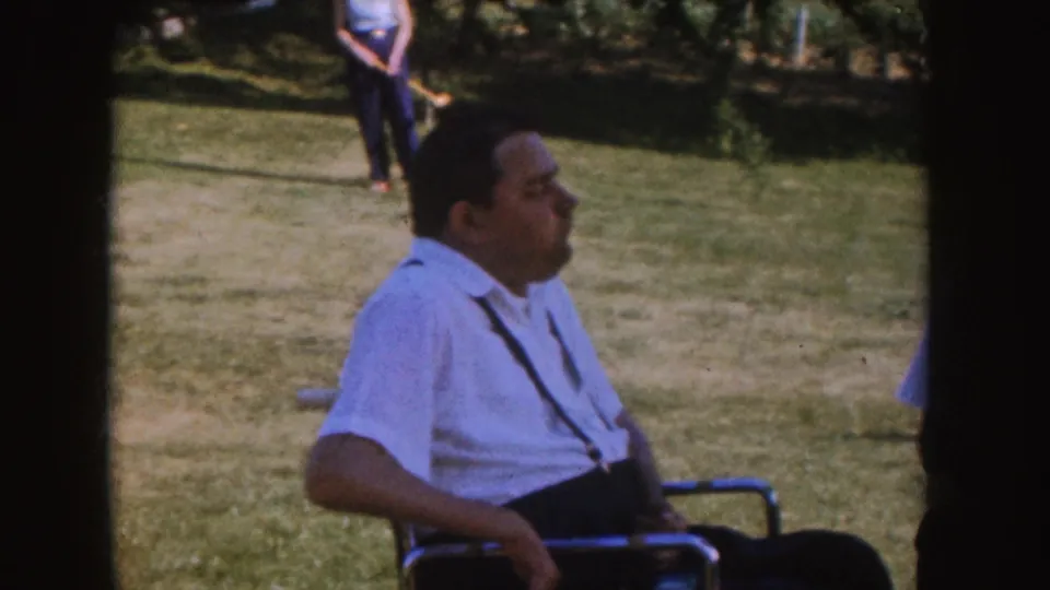 1955 Nebraska home-movie frame showing a man seated in a wheelchair on a backyard lawn, with another person standing behind him.