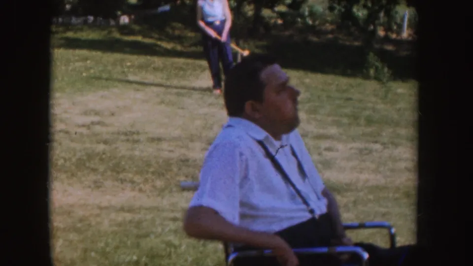 1955 Nebraska backyard picnic frame with a man in a wheelchair seen in profile and a person standing farther back on the lawn.