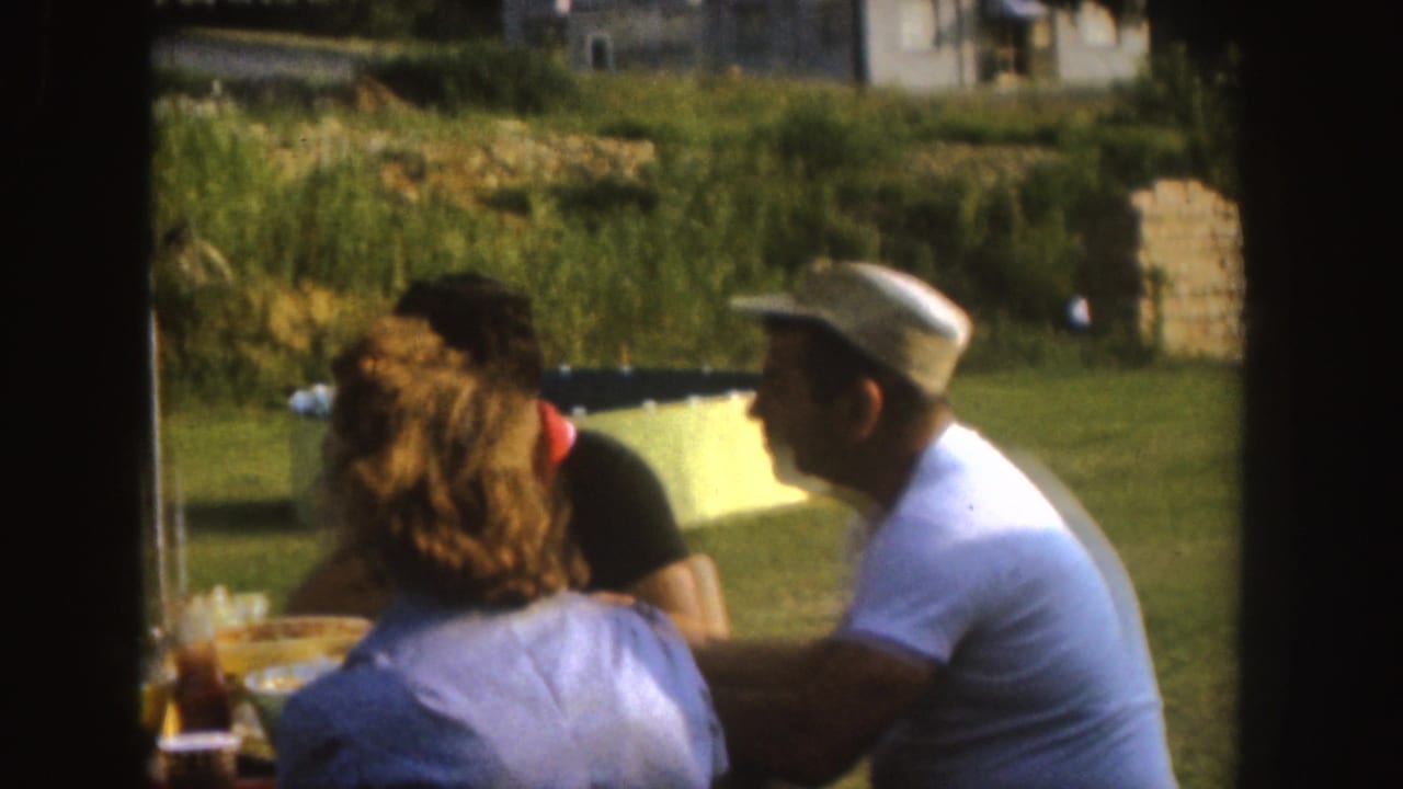 1955 Nebraska home-movie frame with adults seated at a picnic table in a sunny yard, including a man wearing a white cap and shirt.